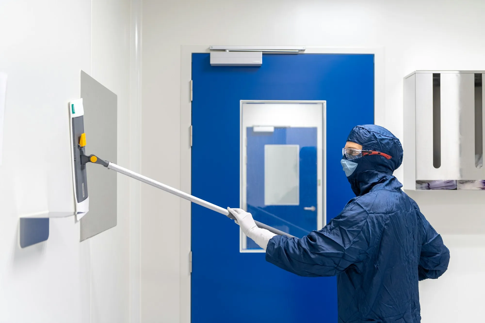 Cleaner in protective gear wiping a door in a white facility